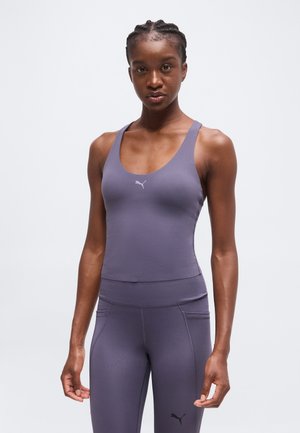 Femme portant un débardeur de sport violet moulant et des leggings avec des logos Puma, debout devant un fond clair uni.