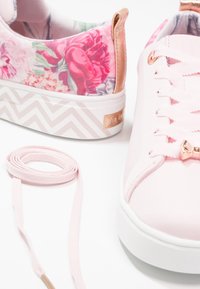Baskets en toile rose avec un motif floral sur le talon, des accents dorés rose texturés, et un motif en zigzag blanc sur la semelle. Elles sont dotées d'un lacet rose.