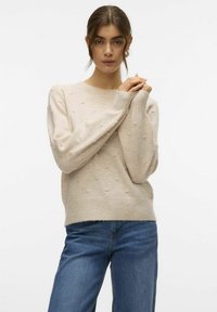 Maglione beige lavorato a maglia con texture a pois in rilievo, scollatura tonda, maniche lunghe, polsini e orlo a costine, abbinato a jeans in denim blu.