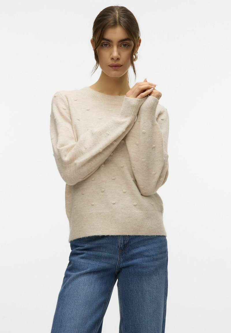 Maglione beige lavorato a maglia con texture a pois in rilievo, scollatura tonda, maniche lunghe, polsini e orlo a costine, abbinato a jeans in denim blu.