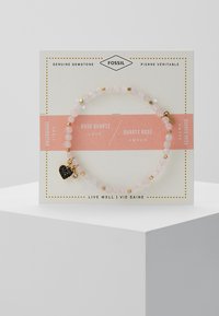 Bracelet en quartz rose avec des accents dorés et un charm cœur noir. Présenté sur une carte blanche avec du texte rose et doré.