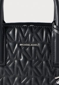 Μαύρη δερμάτινη τσάντα χειρός με καπιτονέ σχέδιο και λογότυπο "MK", ασημένια μεταλλικά στοιχεία και χερούλια στην κορυφή. Το όνομα της μάρκας είναι χαραγμένο μπροστά.