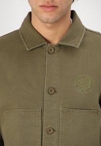 Olive green jacket made of denim, featuring a collared neckline, two chest pockets, brown buttons, and an embroidered logo on the left side.
