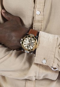 Auksinio tono laikrodis su grietinėlės spalvos ciferblatu ir trimis subdialais, rudas odinis dirželis, sidabro ir aukso detalės, vilkimas ant marškinėlių su atviru kraštu.