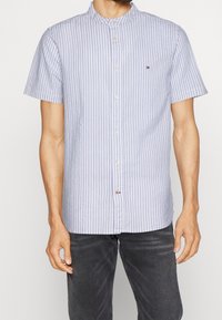 Camisa de manga corta con botones en rayas azul claro y blanco, con cuello mao y un pequeño parche con logo en el pecho.