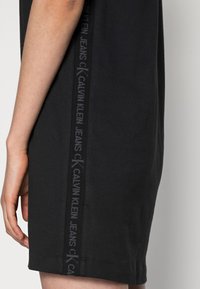 Sidsyn på en person som bär en svart klänning med en vertikal Calvin Klein Jeans-logotypstrimma längs sömmen.