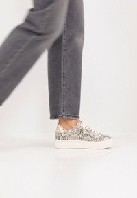 Αθλητικά παπούτσια με λευκή πλατφόρμα, διακοσμημένα με σχέδιο φιδιού σε μπεζ και μαύρο χρώμα, και ανοιχτόχρωμους κορδόνια. Φορεμένα με γκρίζα τζιν.