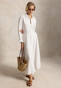 Γυναίκα με μακρύ λευκό φόρεμα με κουμπιά και γυαλιά ηλίου, κρατώντας υφασμένη τσάντα ώμου και φορώντας καφέ σανδάλια, στέκεται μπροστά σε απλό φόντο.