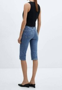 Denimowe capri w średnim niebieskim kolorze, o dopasowanym kroju, skróconej długości, z podwiniętymi nogawkami i tylnymi kieszeniami, w połączeniu z czarną bezrękawnikową bluzką.