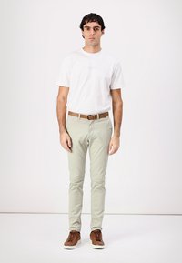 T-shirt bianca in cotone, pantaloni slim-fit verde chiaro, cintura in pelle marrone e sneakers marroni. Modello in piedi su uno sfondo semplice.