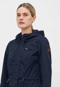 Veste bleu marine avec capuche, dotée d'une poche zippée, de cordons de serrage à la taille, et d'un accent en cuir marron sur l'épaule. Texture lisse.