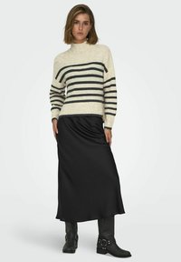 Pull en tricot crème avec des rayures noires, col montant, associé à une jupe maxi satin noire et des bottines noires à boucles.