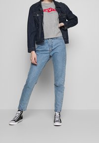 Chaqueta de mezclilla en azul oscuro sobre una camiseta gris con un logo rojo, combinada con vaqueros de cintura alta en azul claro y zapatillas negras de caña alta.