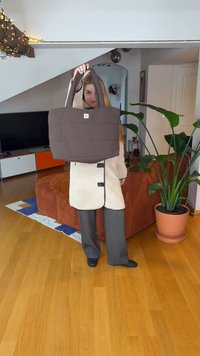 Braune Tragetasche mit zwei stabilen Griffen, ausgestattet mit einem Logo-Patch und einem schlichten, gesteppte Design. Vor einem Hintergrund eines Wohnzimmers platziert.