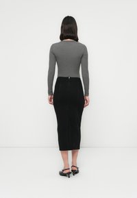 Femeie stând cu spatele, îmbrăcată cu un pulover gri pe termen lung, o fustă midi neagră cu talie înaltă și tocuri slingback negre pe fundal alb.
