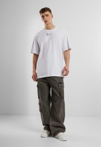 T-shirt en coton blanc à manches courtes avec un petit motif graphique noir sur la poitrine, associé à un pantalon cargo amples gris foncé et des baskets blanches.