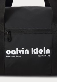 Black nylon duffel bag featuring a zippered pocket and prominent white text branding, including "calvin klein" and "West 39th Street."