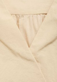 Beige beklædning med en V-formet udskæring og sammensnøring af stoffet ved den øverste brystområde.