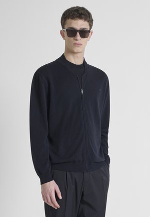 Navy zip-up cardigan made of soft material, featuring a ribbed collar and cuffs, paired with black trousers and dark sunglasses.