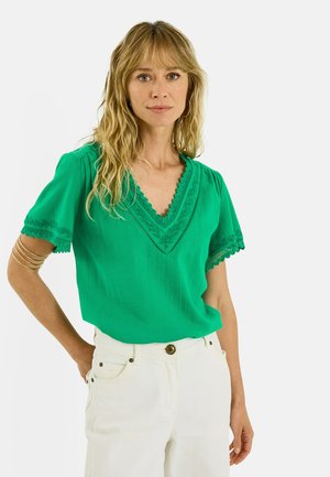 Mujer con cabello largo y rubio que lleva una blusa verde de manga corta con detalles de encaje y pantalones blancos, de pie con una mano en la cadera.