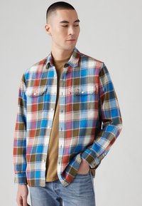 Geruite shirt in blauw, rood, groen en wit. Beschikt over twee borstzakken, knoopsluiting en lange mouwen. Draag het over een beige shirt.