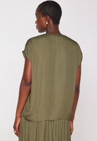 Top verde oliva a maniche corte realizzato in tessuto morbido. Presenta una vestibilità rilassata e un orlo arrotondato, indossato sopra una gonna verde plisse.