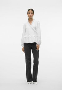 Blusa avvolgente bianca con orlo e maniche a ruffle, abbinata a jeans neri a gamba larga. La modella indossa scarpe con tacco bianche e ha i capelli lisci.