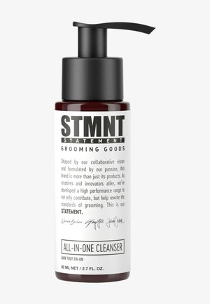 Bouteille en verre rouge avec un bouchon pompe noir. L'étiquette blanche présente un texte noir en gras : « STMNT GROOMING GOODS » et « ALL-IN-ONE CLEANSER ».