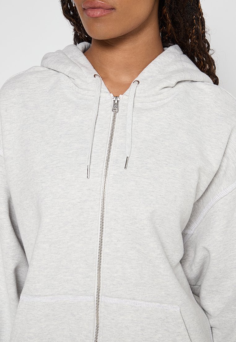 Helles graues Zip-Hoodie aus weichem Material, mit einer Fronttasche, Kordelzügen und gerippten Bündchen. Glatte Textur mit lockerem Sitz.