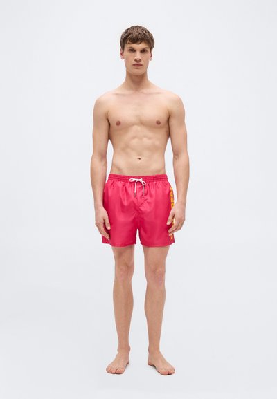 Νέος άνδρας που στέκεται ξυπόλητος φορώντας φωτεινά κόκκινα μαγιό με κίτρινο κείμενο στο πλευρό, μπροστά σε απλό ανοιχτό γκρι φόντο.