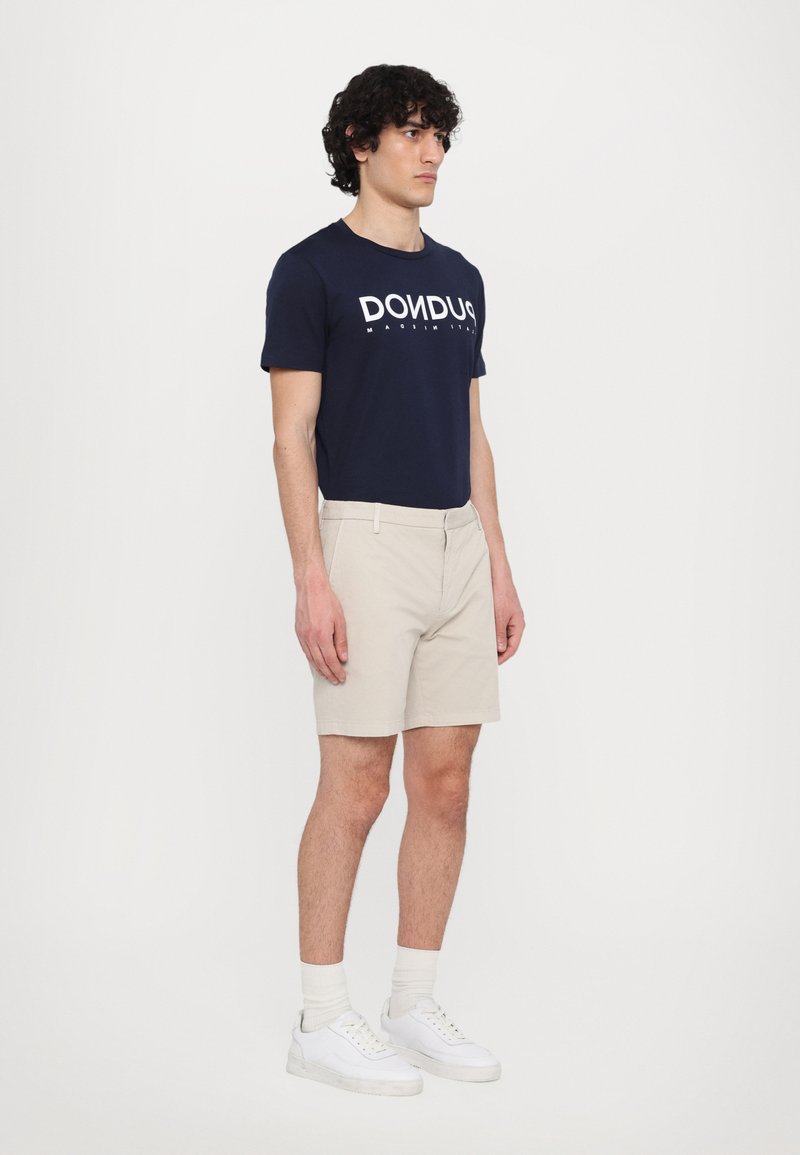 T-shirt blu navy con testo stampato bianco, pantaloni beige e sneakers bianche con suole grigie chiare. Il modello è in posizione laterale contro uno sfondo neutro.