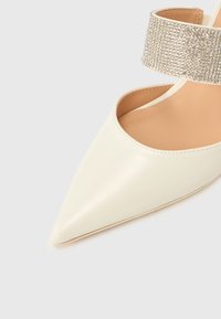 Λευκό δερμάτινο παπούτσι με μυτερή μύτη, ανοιχτές πλευρές και λαμπερό λουράκι στον αστράγαλο. Ομαλή υφή, μινιμαλιστικός σχεδιασμός και κομψό σχήμα.