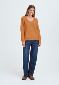 Hoedkleurige gebreide trui met een V-hals, losse pasvorm en lange mouwen, gecombineerd met donkerblauwe rechte jeans en bruine schoenen.