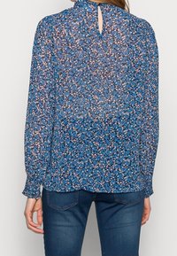 Kvinna i en blå blommig långärmad blus med en liten öppning i ryggen, ihopparad med mörkblå jeans.