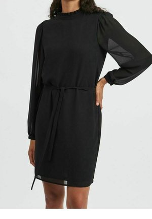 Vrouw draagt een zwarte jurk tot op de knie met doorschijnende lange mouwen, een geruite kraag en een strikriem in de taille, staand met één hand op haar heup.