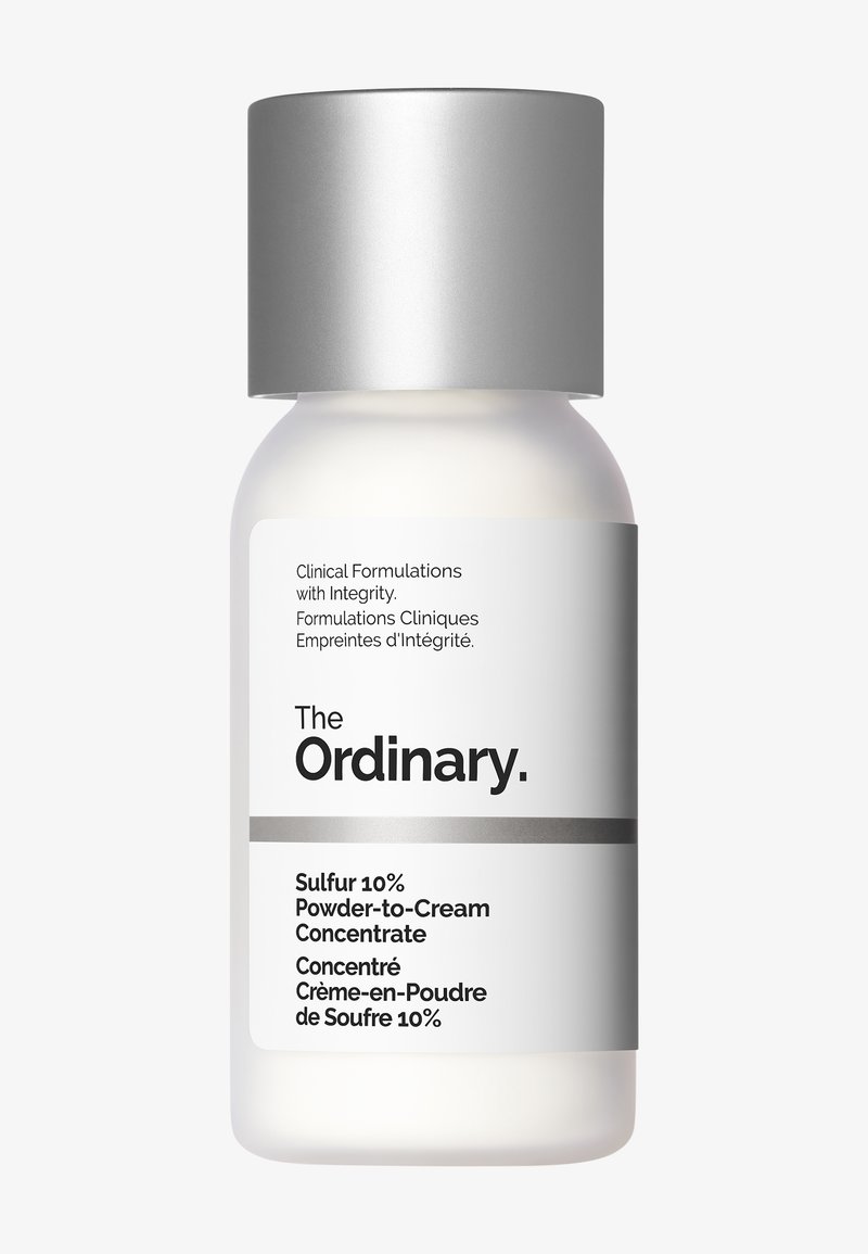 Witte fles met een zilveren dop, gelabeld "The Ordinary," met "Zweefstof 10% Poeder-naar-Crème Concentraat." Gladde textuur, minimalistisch ontwerp.