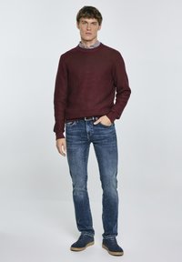 Bordeauxfarbener ribbeliger Strickpullover, blaue Jeans und dunkelblaue Schuhe. Das Model posiert mit den Händen auf den Hüften und trägt ein Hemd mit Kragen darunter.