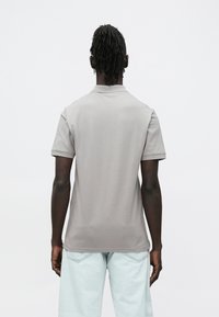 Šedé polo tričko s golierom, krátkymi rukávmi a hladkým materiálom, nosené s priliehavými svetlomodrými nohavicami voľného strihu. Zobrazený je pohľad zozadu.