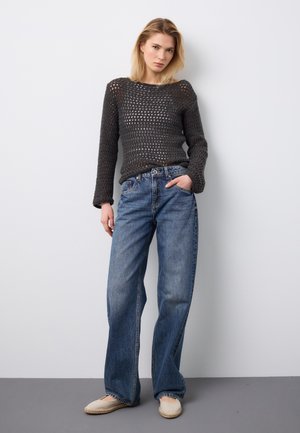 Jonge vrouw staande tegen een witte muur, draagt een zwarte gehaakte trui, losse blauwe spijkerbroek en beige instapschoenen, met één hand in de zak.