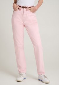 Jeans rosa chiaro a vita alta con tasche laterali, leggera trama e cintura marrone. Abbinati a scarpe sportive bianche.