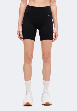 Personne portant un short de sport noir, des chaussettes blanches hautes et des chaussures de course blanches, debout devant un fond uni.