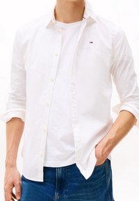 Camicia bianca con maniche arrotolate sopra una t-shirt bianca. Logo ben visibile sul petto, abbinata a jeans in denim blu. Tessuto morbido.