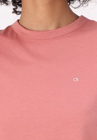 Trumpas rankovėmis, apvaliu kaklu marškinėliai iš minkštos rožinės medvilnės, su mažu baltu "ck" logotipu kairėje krūtinės pusėje. Lygi tekstūra.