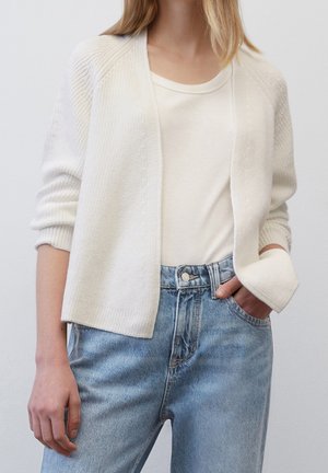 Femme portant un jean taille haute bleu clair, un T-shirt blanc et un cardigan tricoté ouvert écru, avec une main dans la poche et une posture détendue.