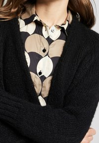 Czarny dziany kardigan o fakturowanym wykończeniu, noszony na wzorzystej koszuli z dużymi beżowymi i czarnymi kołami oraz czarnymi guzikami.