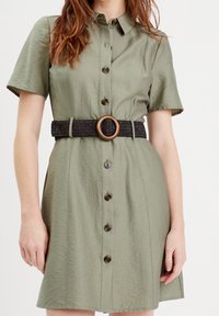 Robe chemise courte à manches courtes de couleur olive avec un devant à boutons et une taille ceinturée noire avec une boucle circulaire en bois. Texture de tissu lisse.