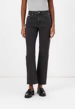 Personne portant un jean noir taille haute à jambes droites, un haut gris sans manches et des mocassins noirs brillants sur un fond blanc uni.