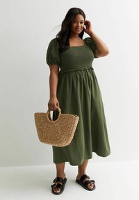 Vestido de manga abullonada en verde oliva con fruncido en el corpino, acompañado de un bolso de paja trenzada y sandalias negras de tiras. Textura de tela suave.