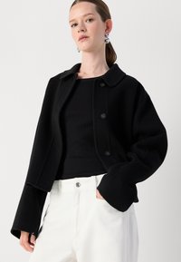 Zwarte cropped jacket met een puntige kraag, knoopsluitingen en brede mouwen, gecombineerd met een zwarte geribbelde top en witte broeken.