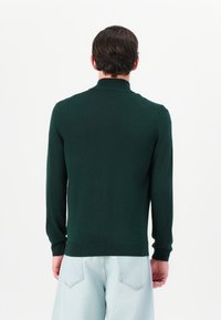 Tumši zaļš adīts džemperis ar augstu apkakli, ribotām aprocēm un jostu, kas valkāts ar gaiši ziliem džinsa biksēm. Nav redzami raksti vai akcenti.